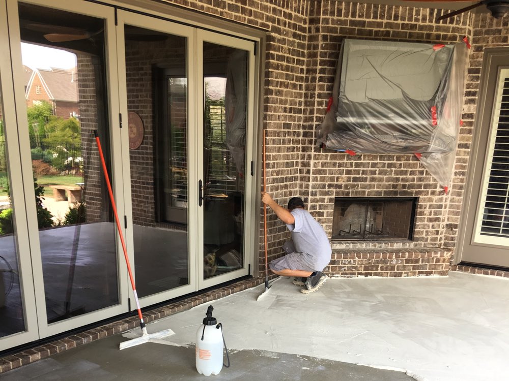 A covered brick porch in Thompson's Station being prepared for staining, the outdoor fireplace masked off in plastic and a pump sprayer standing on the damp slab